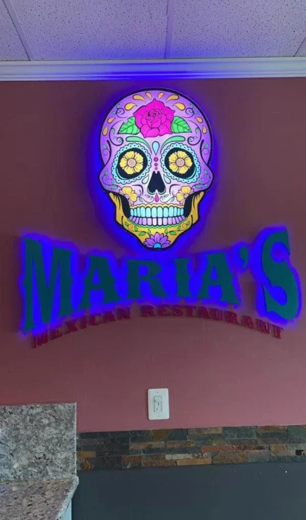 Neon skull sign above “MARIA’S” restaurant on a maroon wall, glowing purple and blue.
