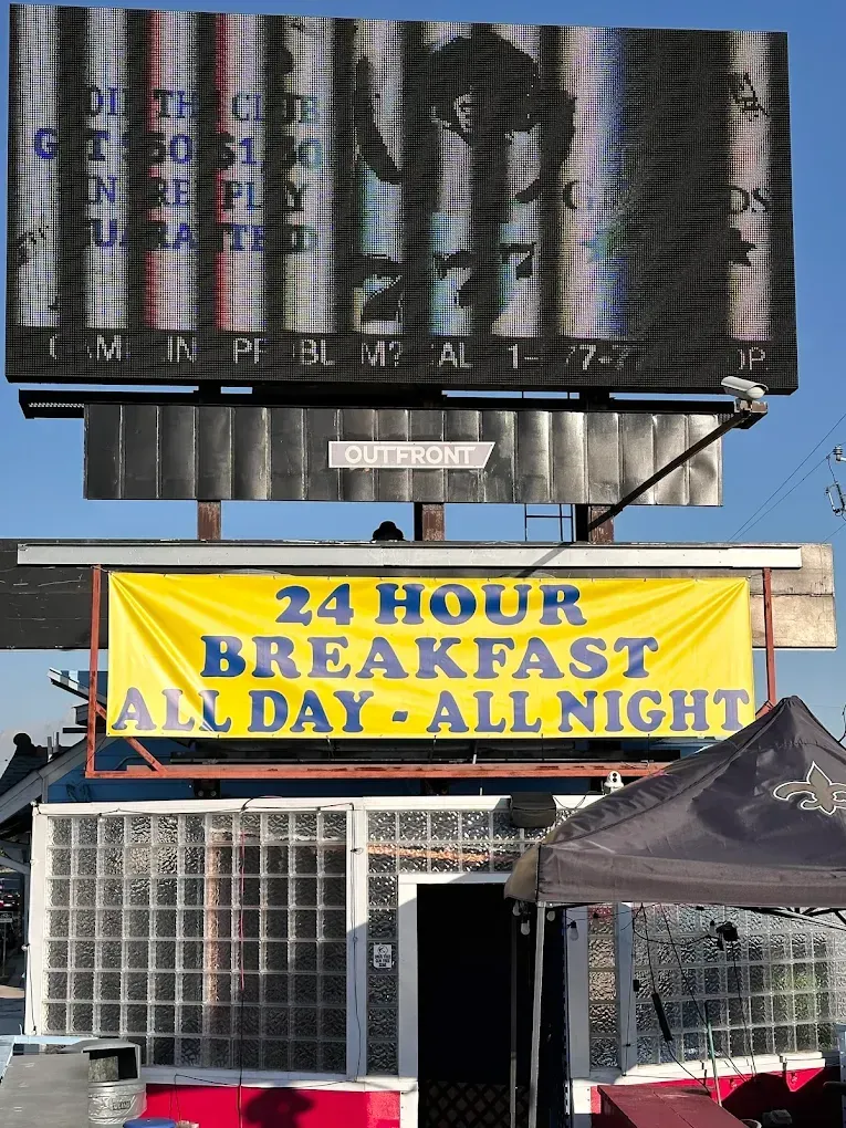 Storefront with a yellow “24 HOUR BREAKFAST ALL DAY! ALL NIGHT!” sign under a billboard.