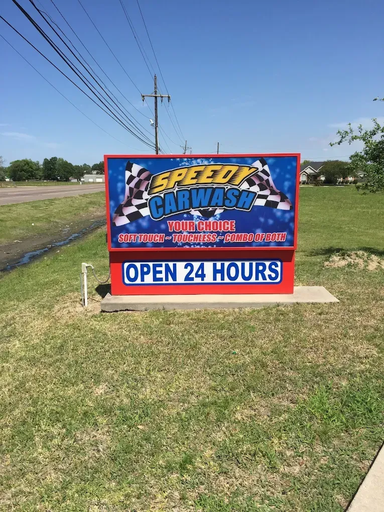 Speedy Car Wash sign in red, blue, and white, with “Open 24 Hours” on a grassy roadside.