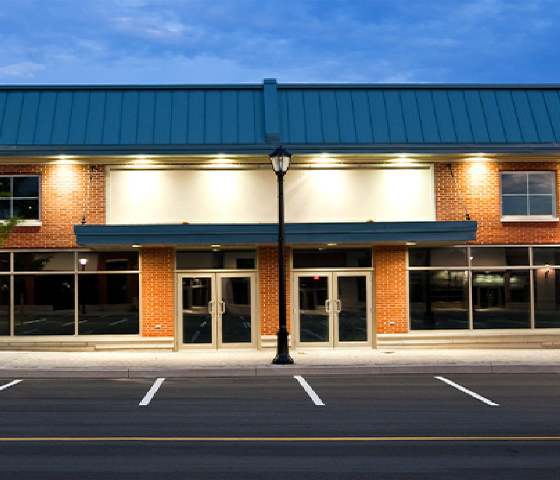 Brick storefront at dusk with teal awning, lit windows, and empty parking spaces in front