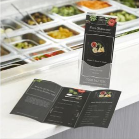 Black restaurant menu standing on a counter beside folded menus and food trays in the background