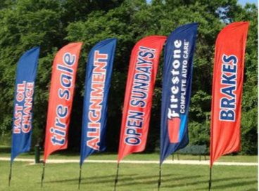 Six colorful roadside advertising flags for tire service and brakes on a grassy field under trees.