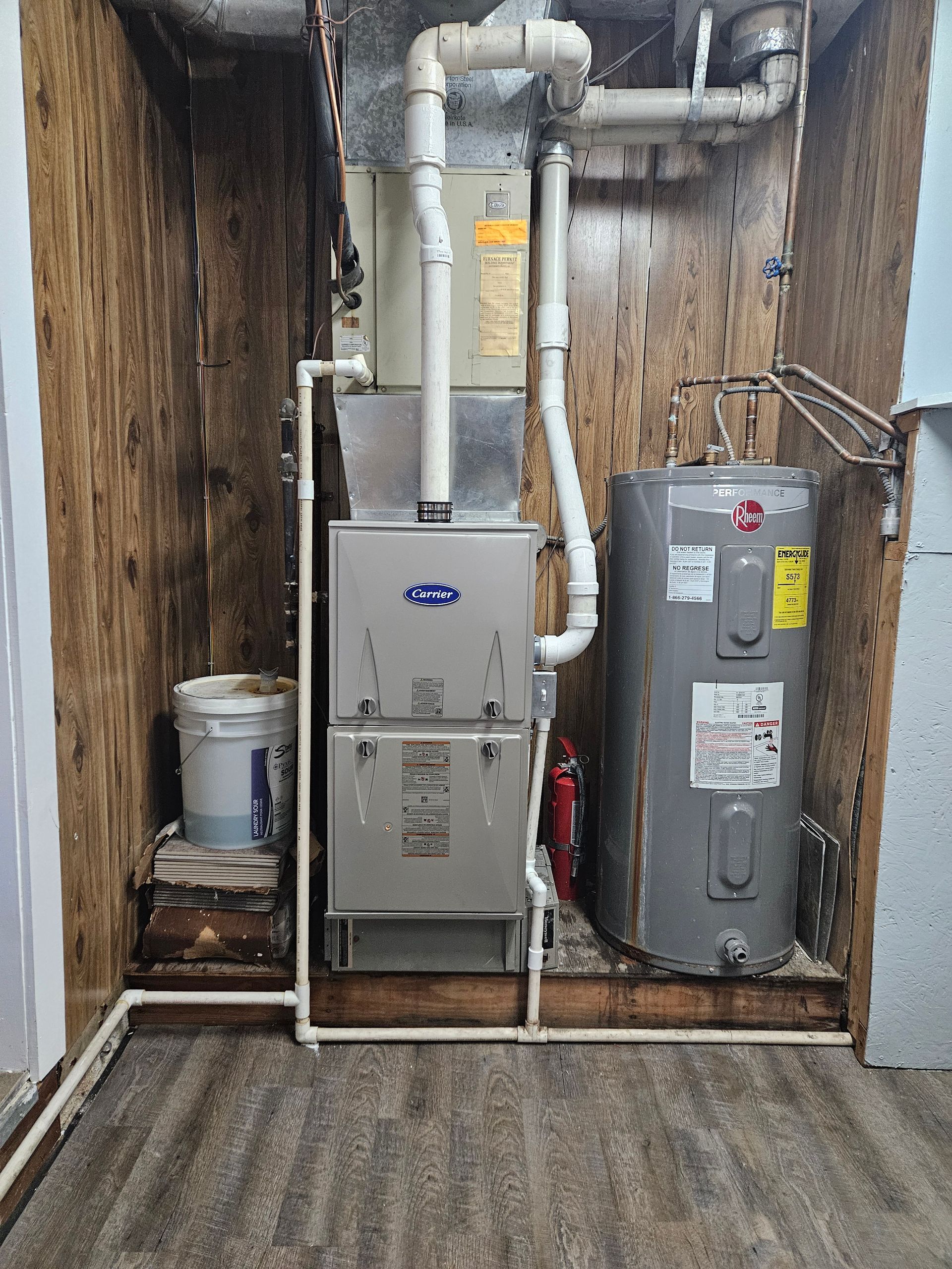 Furnace and water heater in a utility room with wood paneling and a bucket.