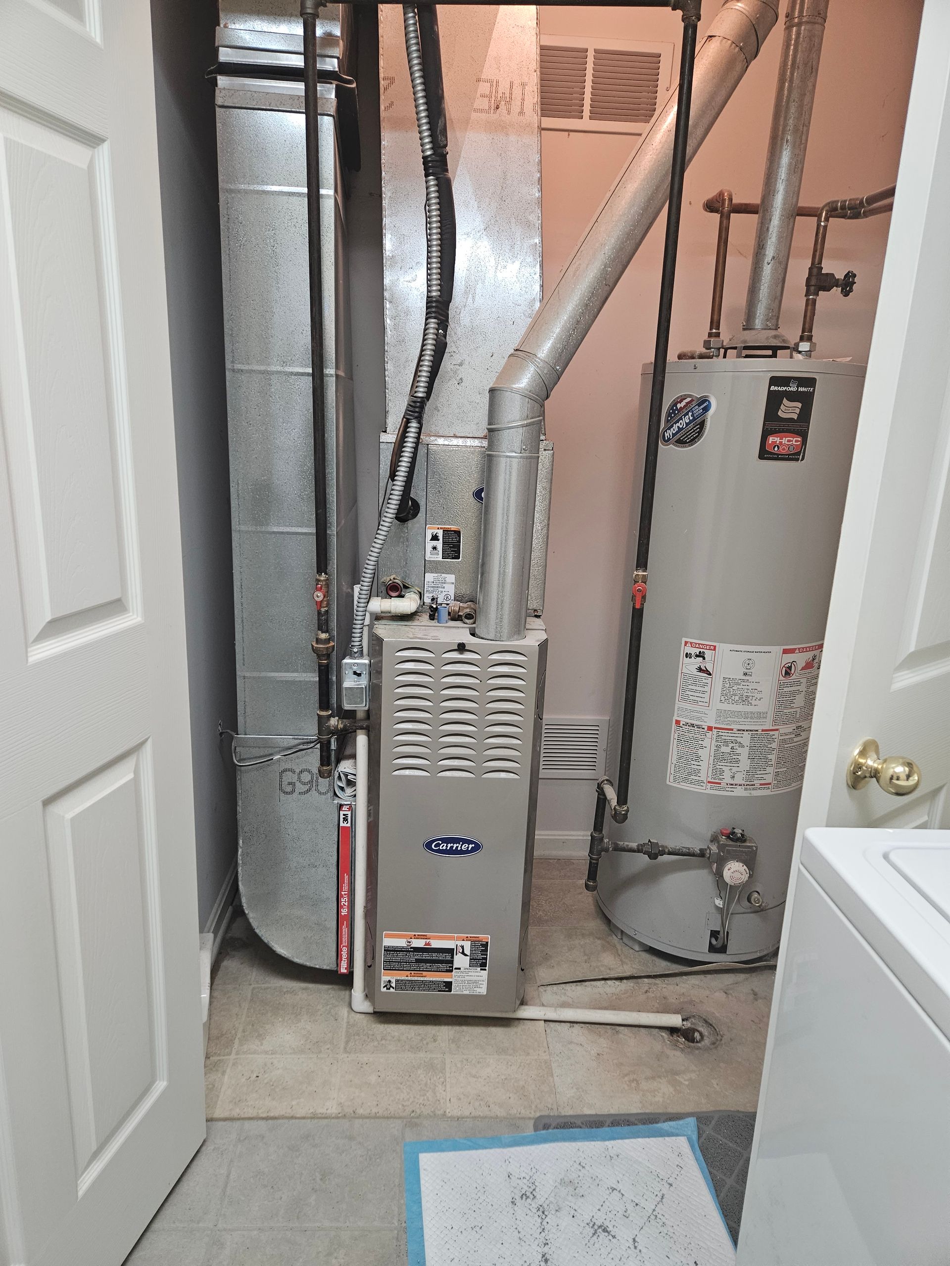 A utility room with a furnace, water heater, and ductwork. Open door on the left; white washing machine on the right.