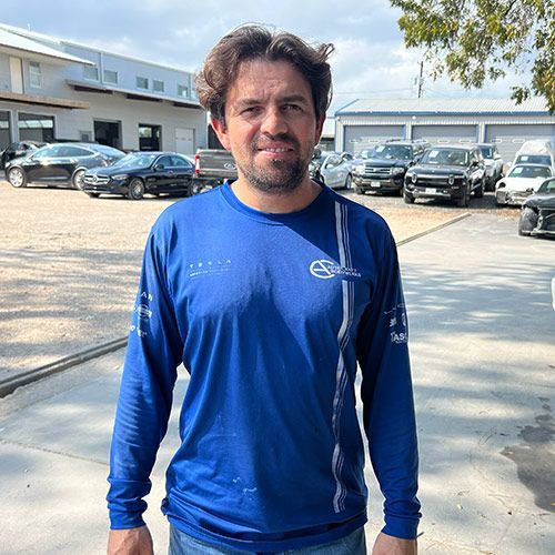 Man in blue long-sleeve shirt stands outside near cars and a building, looking at the camera.