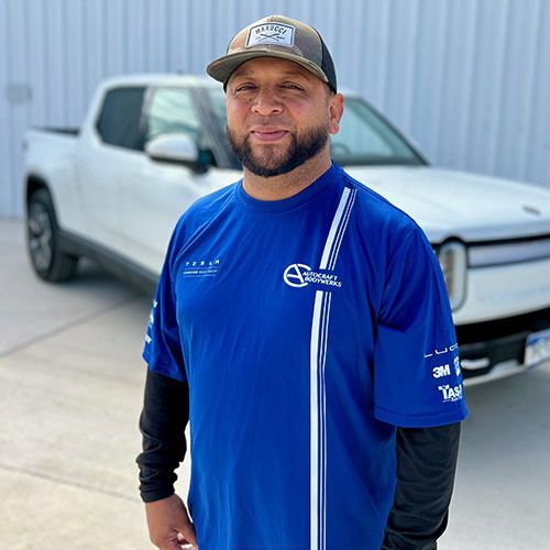 Man with a beard in a black shirt, smiling, in front of a Tesla sign.