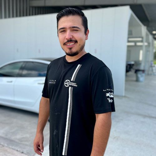 Man in black shirt with logo, smiling in front of white car and building.