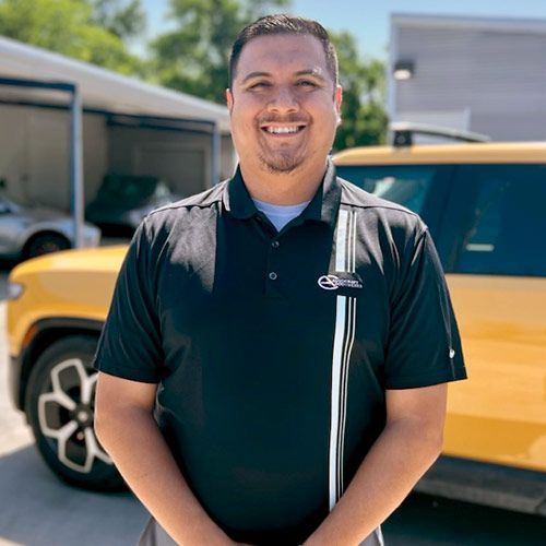 Man smiling, wearing a black polo shirt, standing in front of a yellow car.
