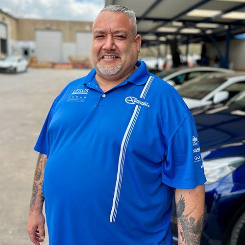 Man in blue shirt with tattoos poses next to cars in an outdoor lot.