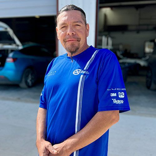 Man in blue shirt, standing outside a garage with a blue car visible in the background.