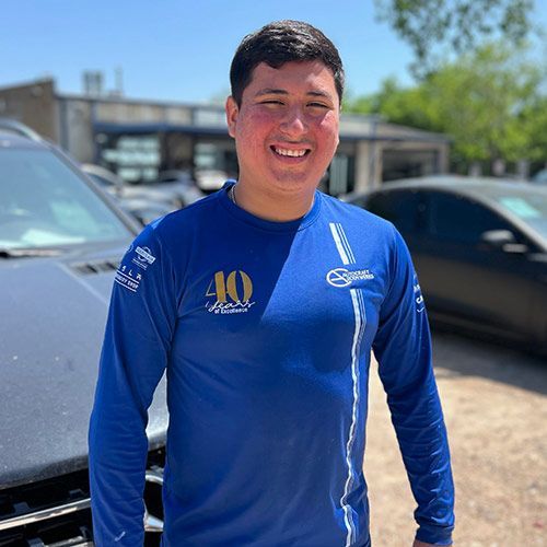 Man wearing a blue shirt smiles outdoors near cars.