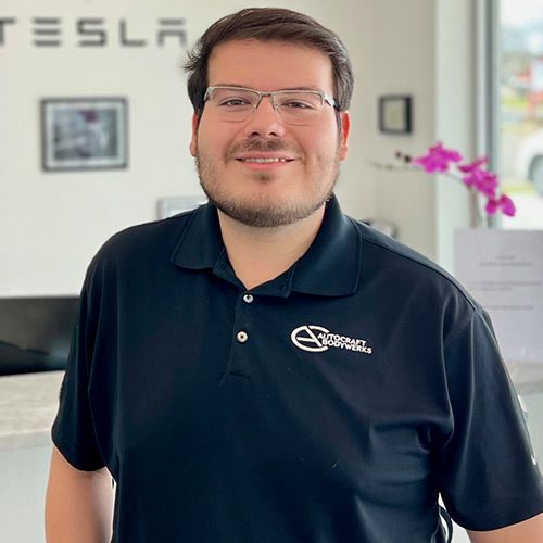 Man wearing glasses and a black polo shirt smiles in front of a Tesla dealership.