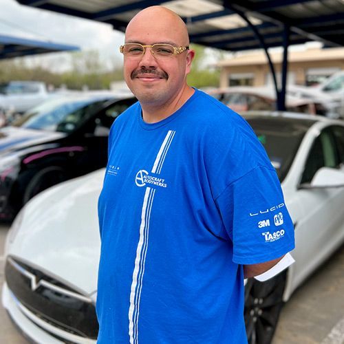 Bald man with glasses smiles, standing in front of a white car. He wears a blue shirt and stands outdoors.