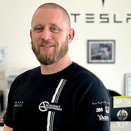 Man with a beard in a black shirt, smiling, in front of a Tesla sign.