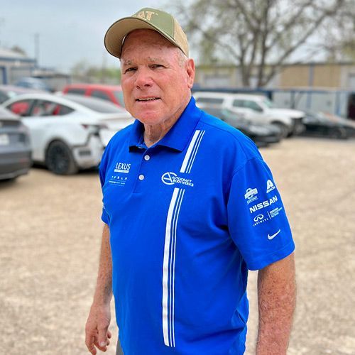Man in blue shirt and cap, standing outdoors near cars; smiling.