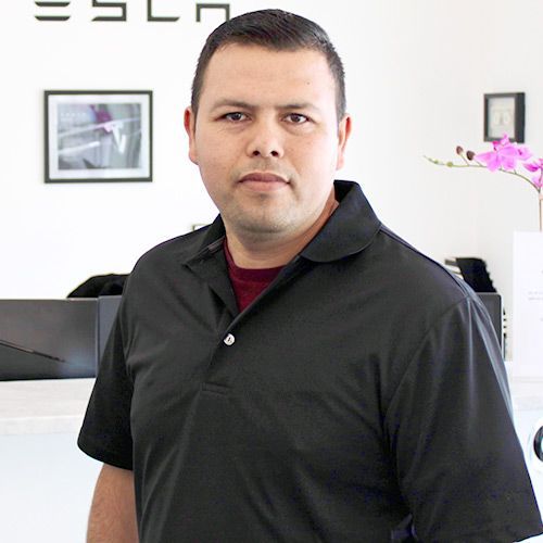 Man in a black polo shirt, standing indoors with a Tesla sign in the background.