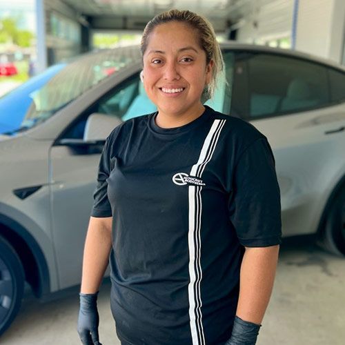 Woman in black shirt and gloves smiles in front of a gray car.