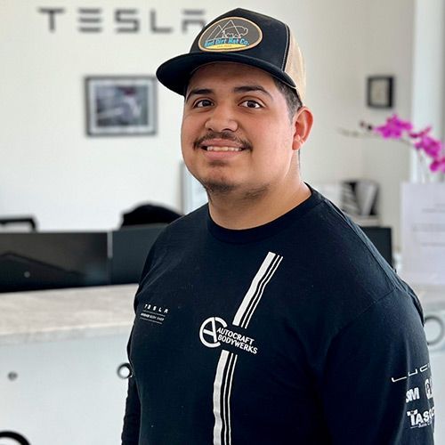 Man in a black shirt and baseball cap smiling, standing in front of a Tesla logo.