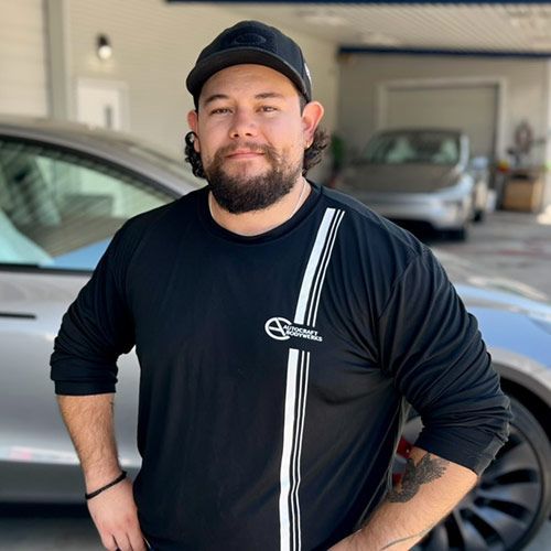 Man in black shirt and cap stands in front of a silver car, smiling.
