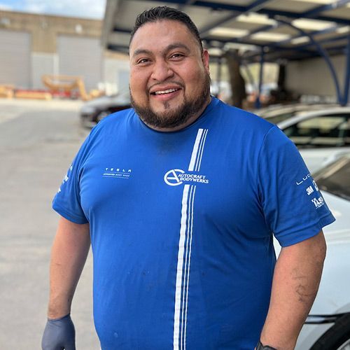 Man in blue shirt smiles, standing outdoors near cars; white stripe on shirt, wearing gloves.