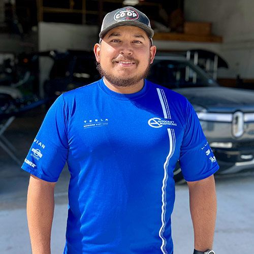 Man in a blue shirt and baseball cap smiles in front of a car. Garage setting.