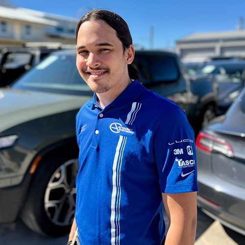 Man in blue polo shirt stands outdoors near cars, smiling.