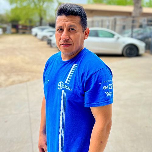 Man in blue shirt, standing outside near parked cars, looking at the camera.
