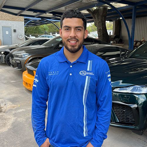Man in blue shirt, smiling, standing in front of several cars at an outdoor shop.