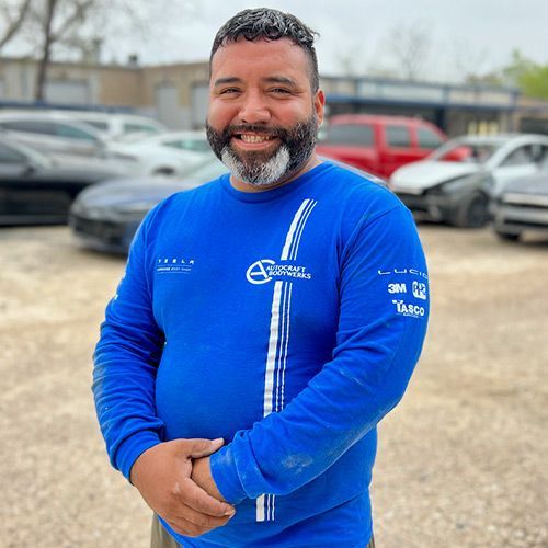 Man in blue long-sleeve shirt smiling, arms crossed. Cars in a lot behind him.