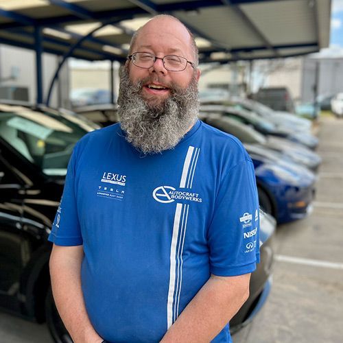 Man with glasses and a gray beard wearing a blue shirt stands in front of a row of cars.