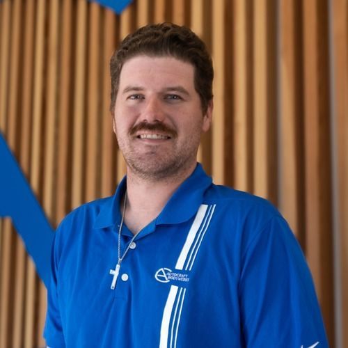Man in a blue polo shirt standing indoors, smiling in front of wood slats and blue accents