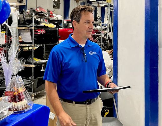 Man in blue shirt, holding clipboard, speaks indoors at a possible celebration.