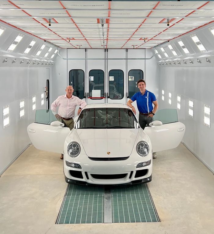 Two men pose with a white Porsche in a paint booth. Both men lean on the car with open doors.