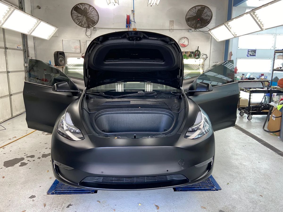 Black Tesla Model Y with hood open, doors ajar, in a garage.