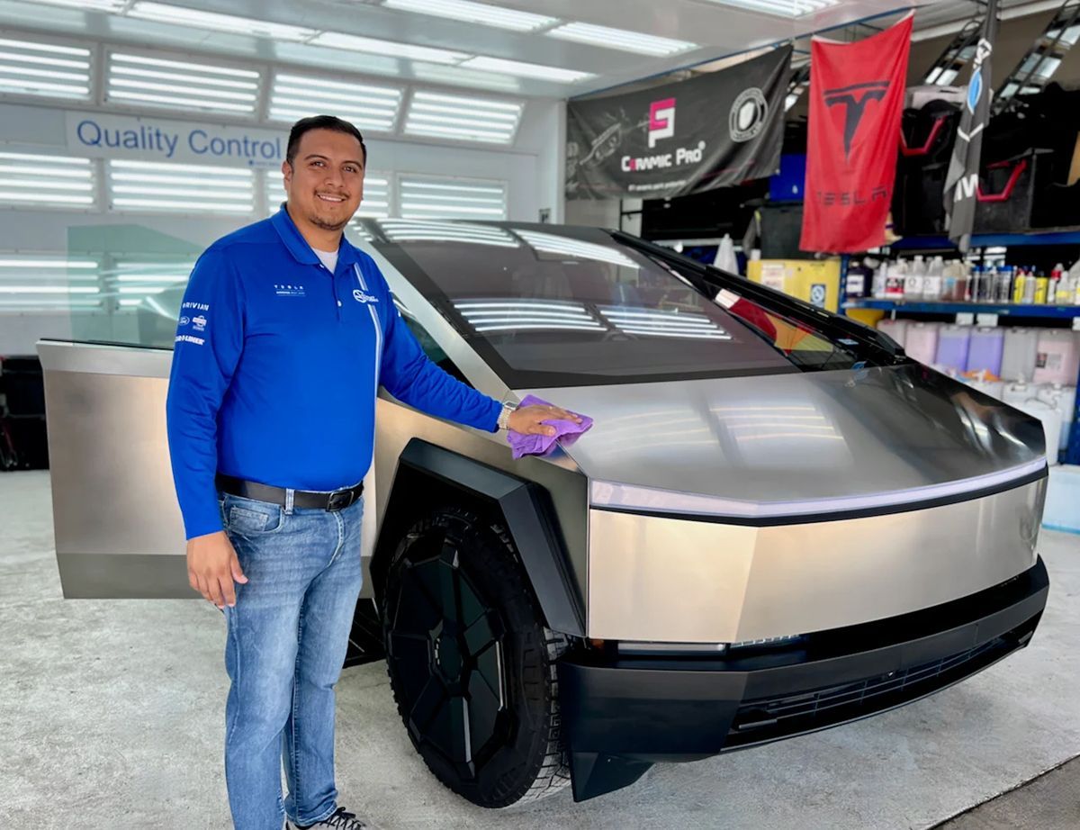 Man in blue shirt wiping a Tesla Cybertruck in a shop.