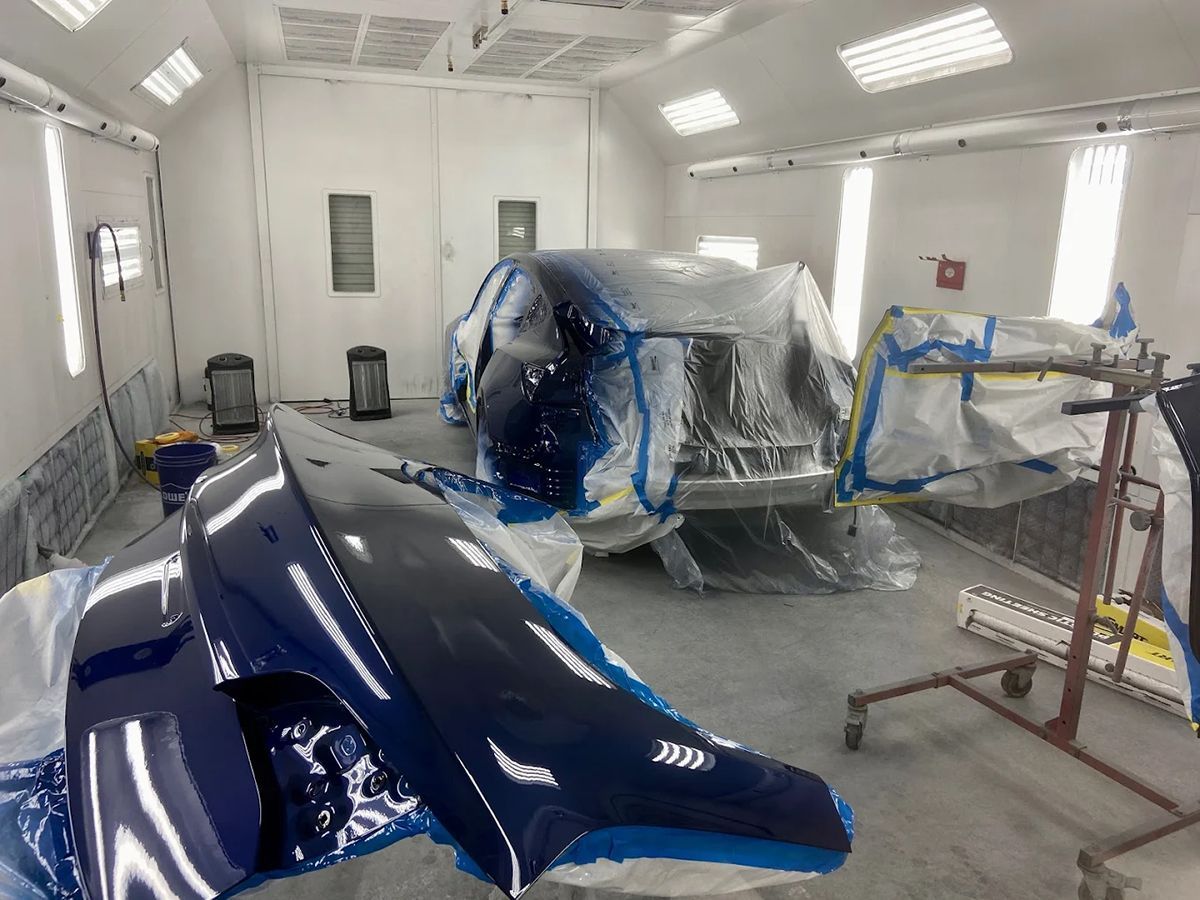 Car in a paint booth, covered in plastic, with painted blue body panels in foreground.