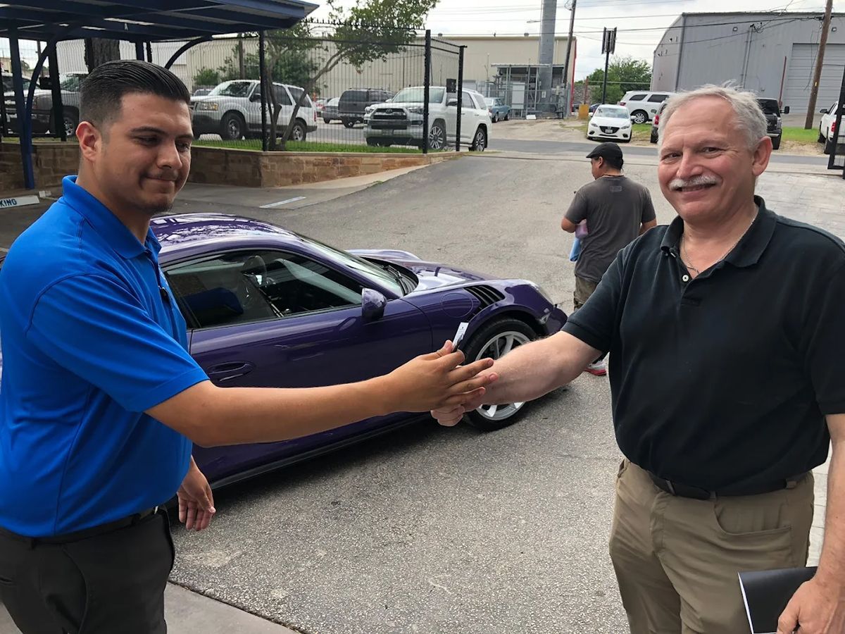 Two men shaking hands beside a purple sports car in an outdoor setting.