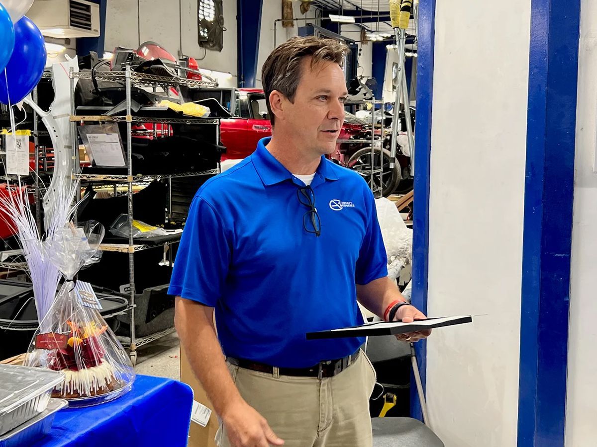 Man in blue shirt speaks, holding a clipboard, indoors. Blue and white decorations are visible.