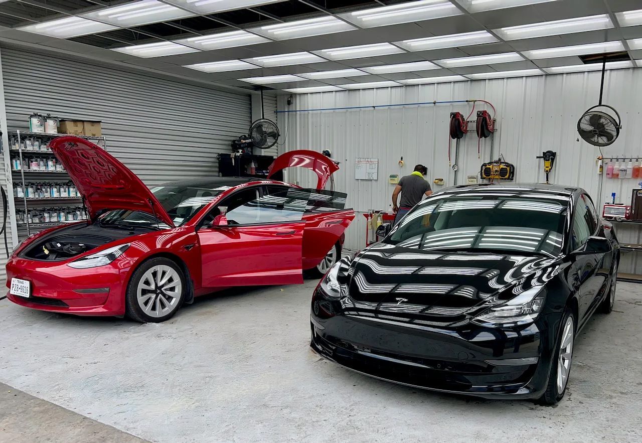 Two cars, red and black, in a repair shop with open hoods. A person works on the black car.