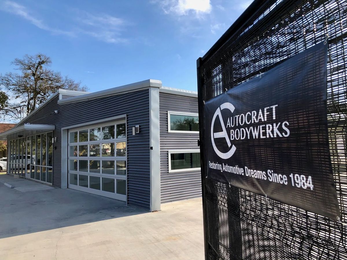 Autocraft Bodywerks building with a banner on a fence. Blue siding, glass garage door, clear sky.