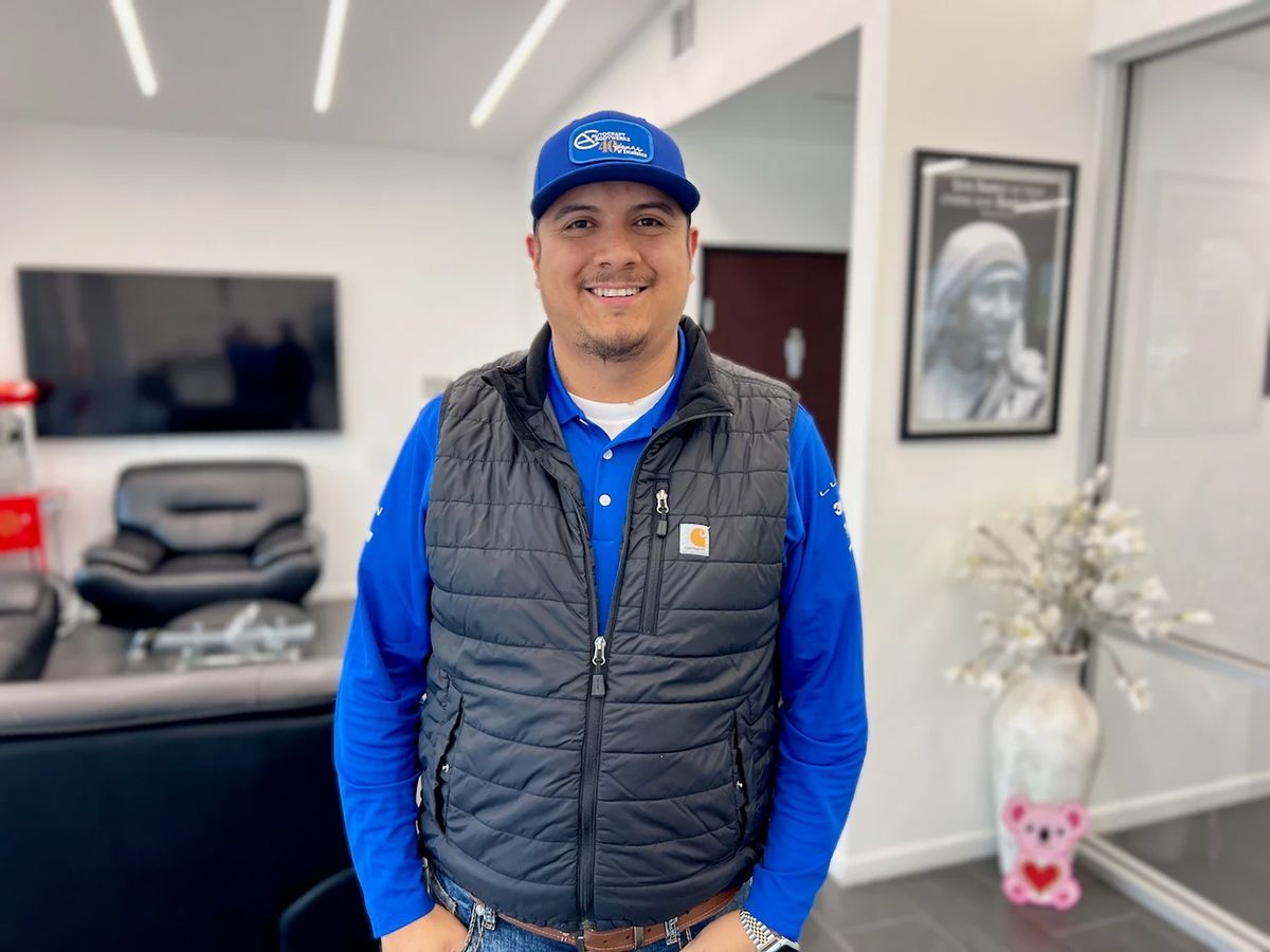 Man in blue shirt, vest, and cap smiling indoors, near artwork and seating.