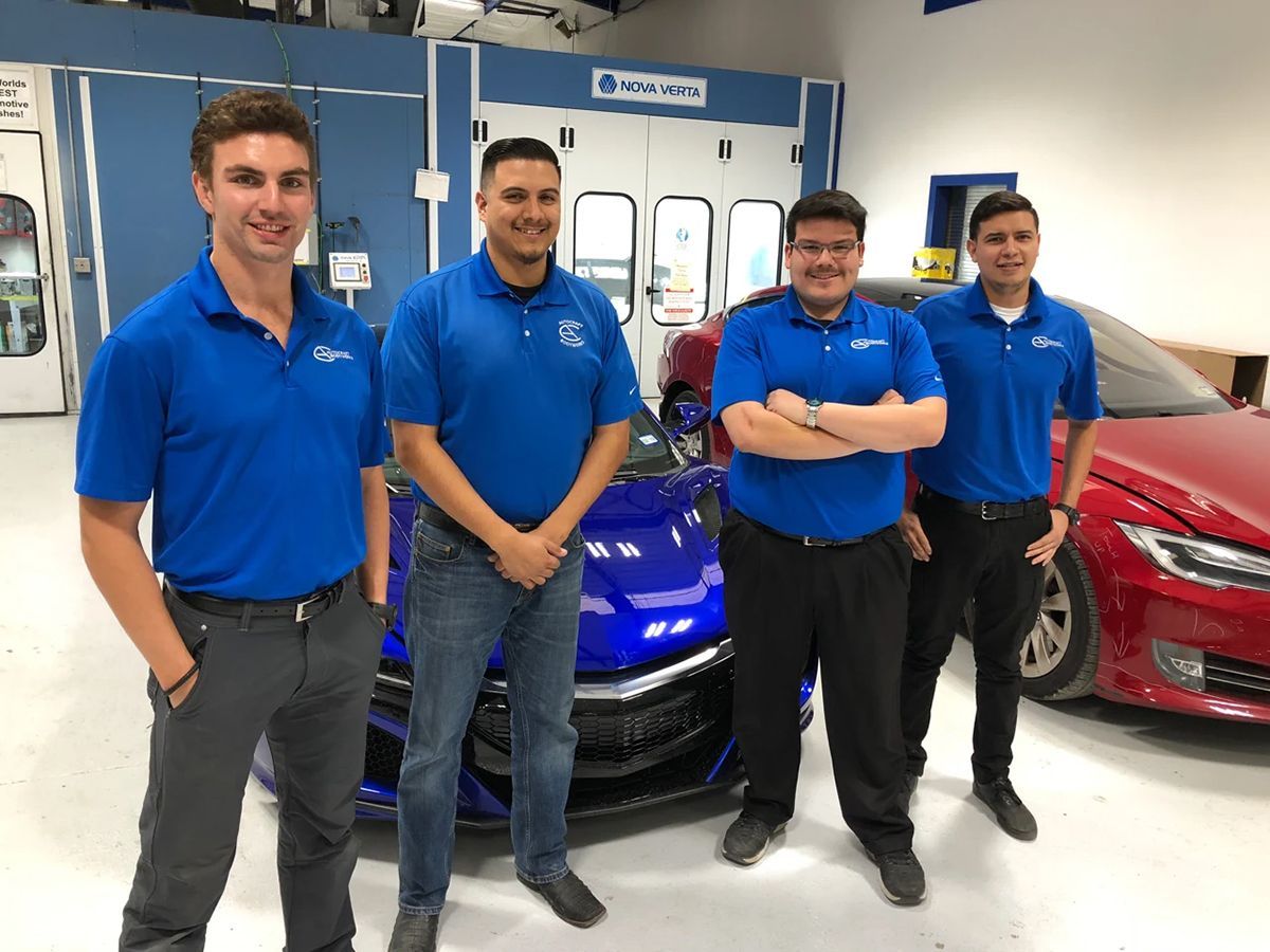 Four people in blue shirts stand in front of cars in a shop.