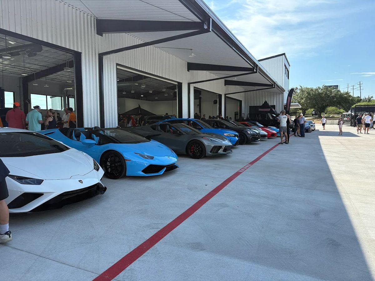 Row of luxury cars parked outside a white building with open garage doors; people gather.