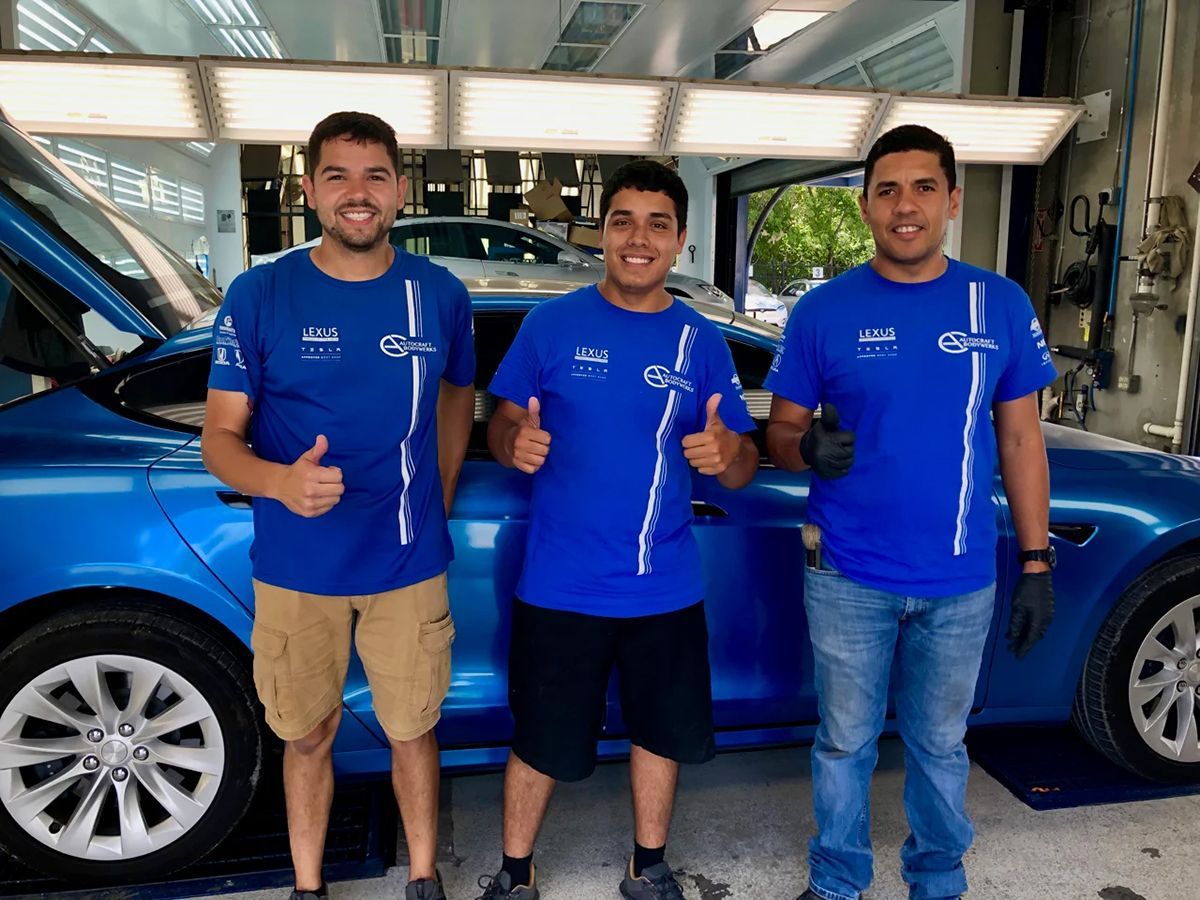 Three men in blue shirts give thumbs-up, standing in front of a blue Tesla with open hood in a workshop.