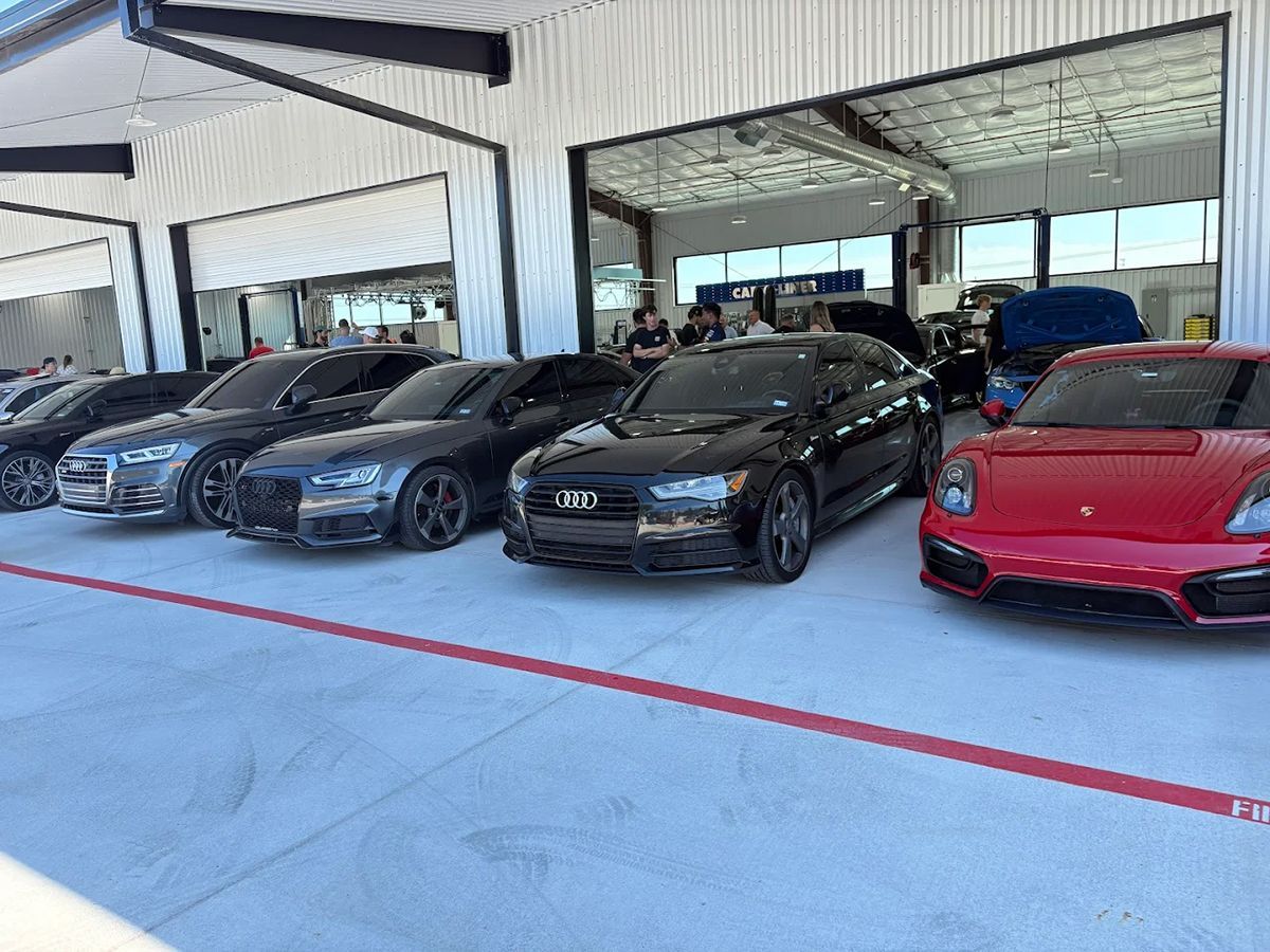 Row of luxury cars parked outside a garage: Audi, Porsche, various colors.
