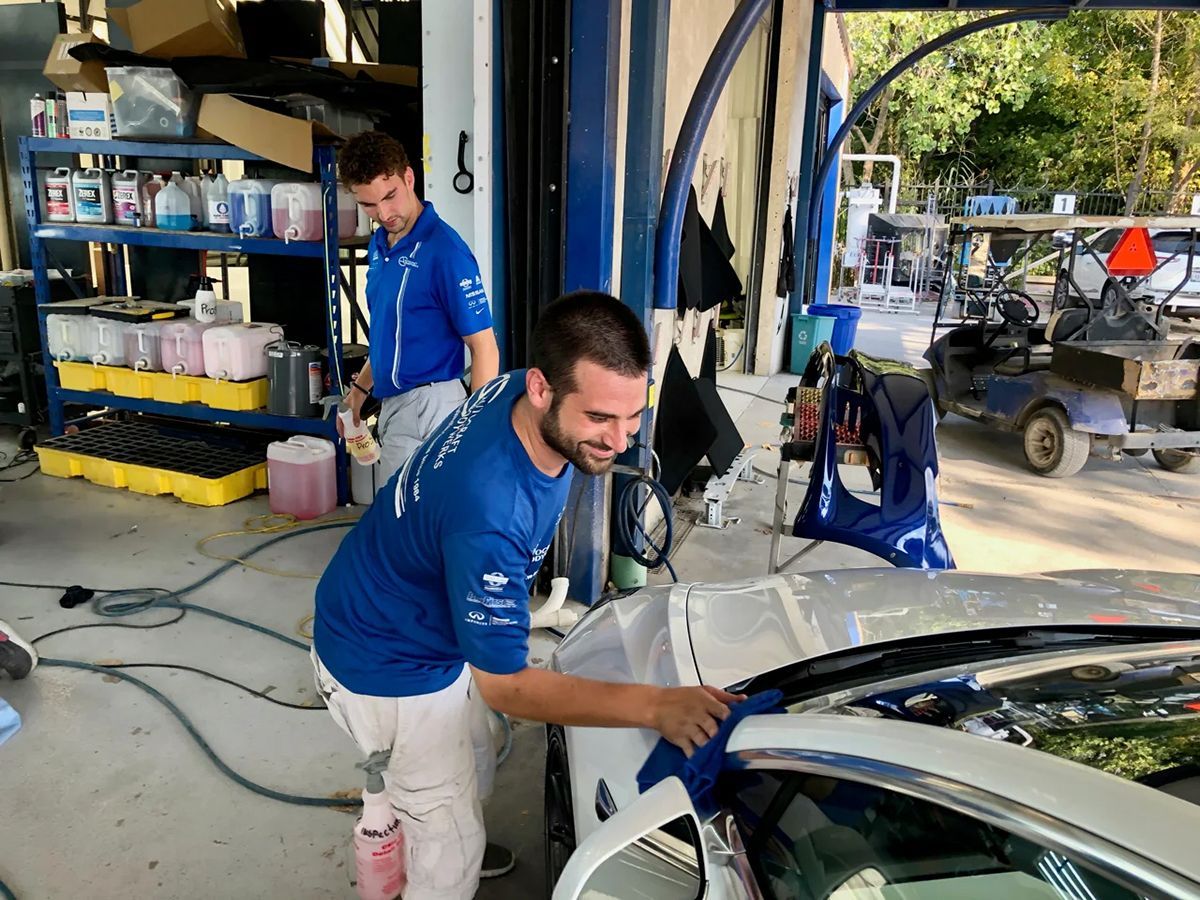 Two men in blue shirts cleaning a white car in an outdoor garage setting.