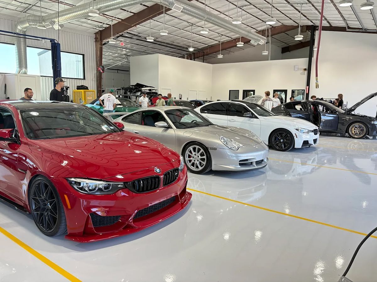 Red BMW, silver Porsche, and other cars in a bright garage with people milling around.