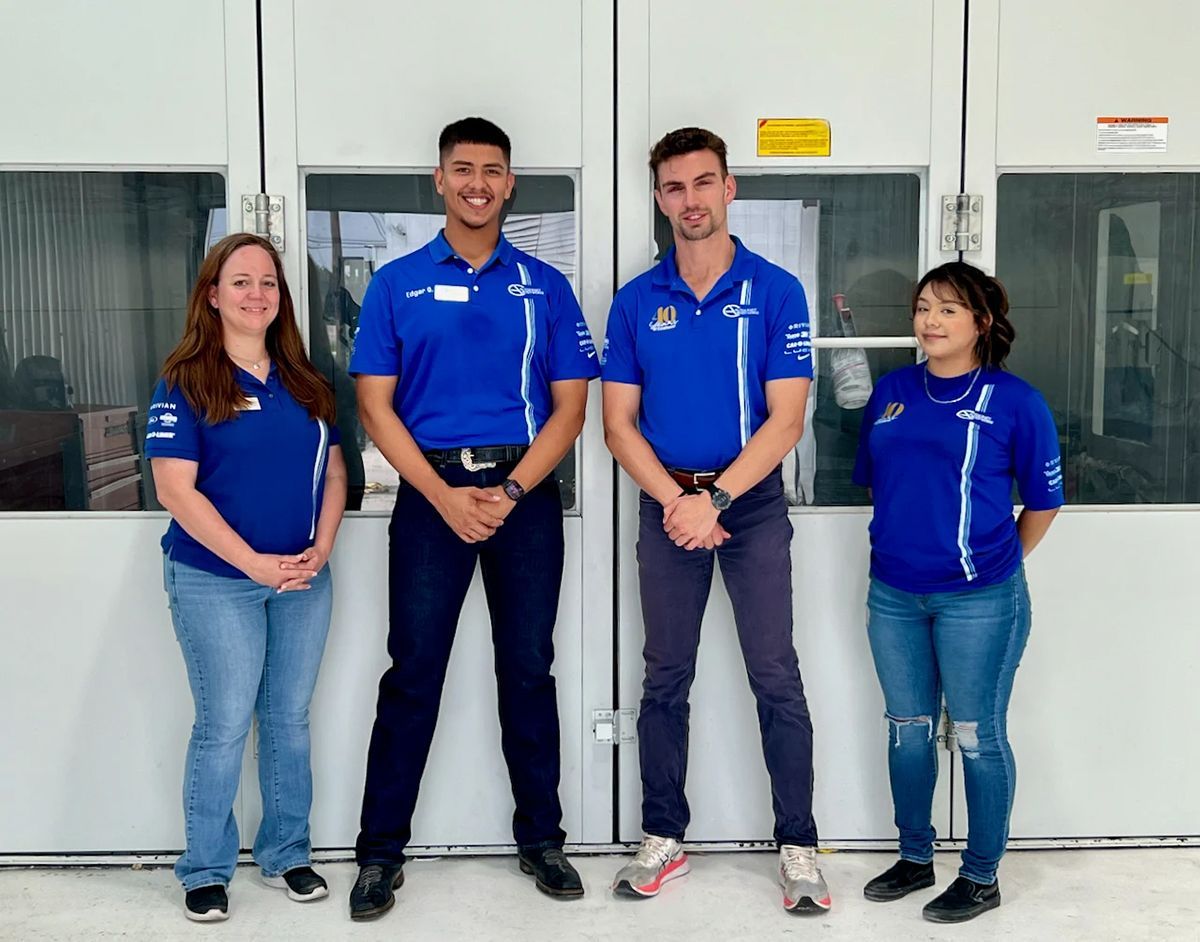 Four people in blue shirts and jeans stand in front of industrial doors, smiling.