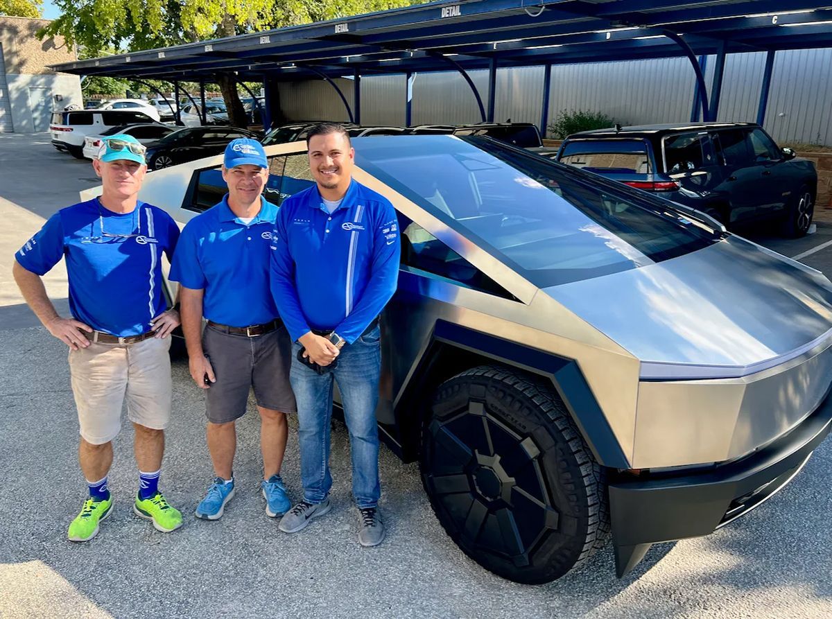 Three men in blue shirts pose with a silver Tesla Cybertruck outside on a sunny day.