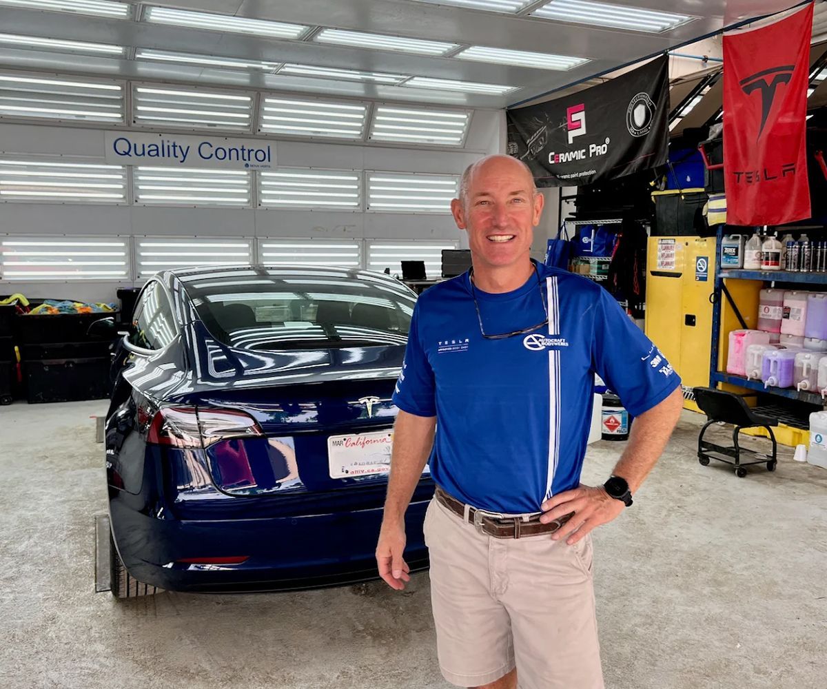 Man standing in a garage with a blue Tesla, wearing a blue shirt and khaki shorts.